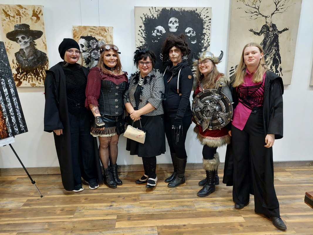 Six people dressed up in steam punk gothic attire posing for the camera while attending an art show by local alternative/gothic artist Ethan Thom. 