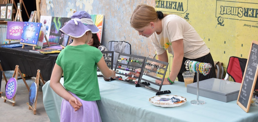 Two young girls interacting at an art booth. One is selling jewelry and the other is browsing. 